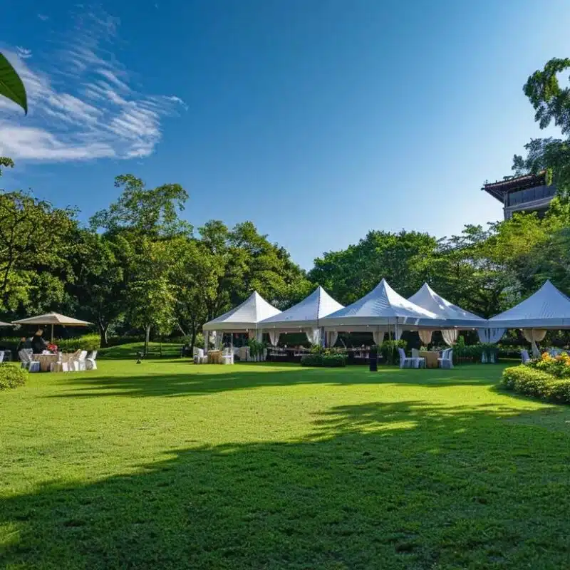 a vibrant outdoor event scene showcases elegantly arranged tents surrounded by lush greenery, contrasting with a distant traditional venue under a clear blue sky, capturing the essence of celebration in a natural setting.