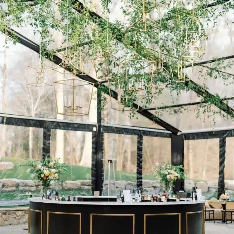 A sleek, circular black and gold bar with floral arrangements, set under a clear event tent with modern hanging decor.
