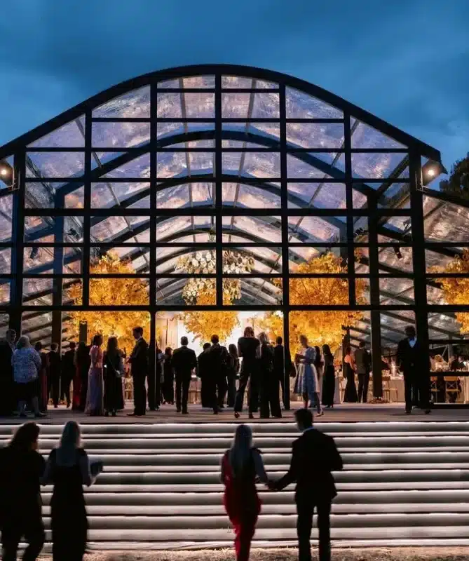 Grand glass-structured event tent at night, illuminated with warm lighting, hosting a formal corporate gathering.