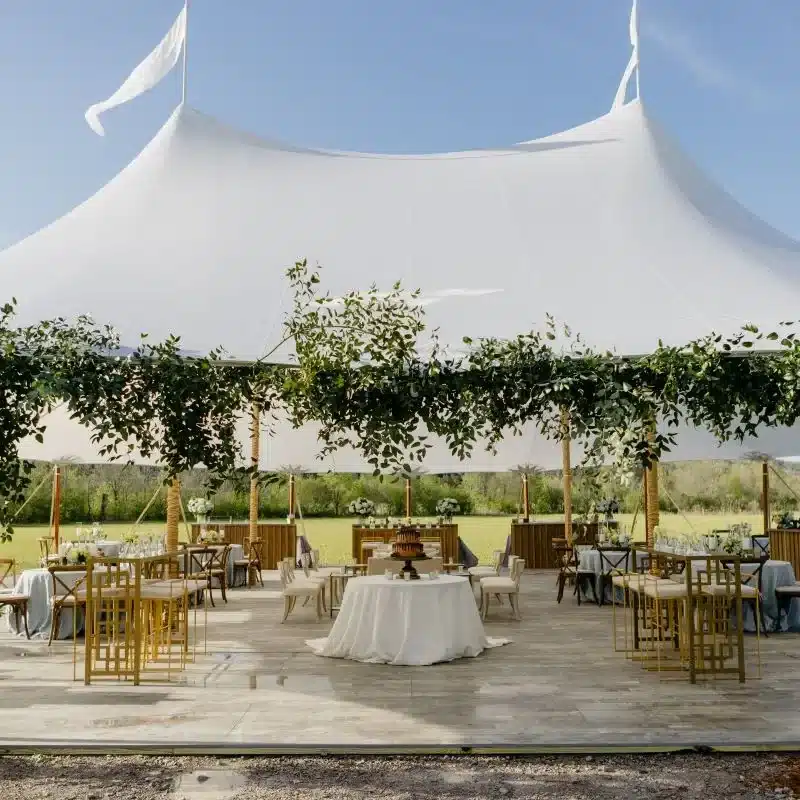 Outdoor event with a white sailcloth tent, tables, and greenery decorations.