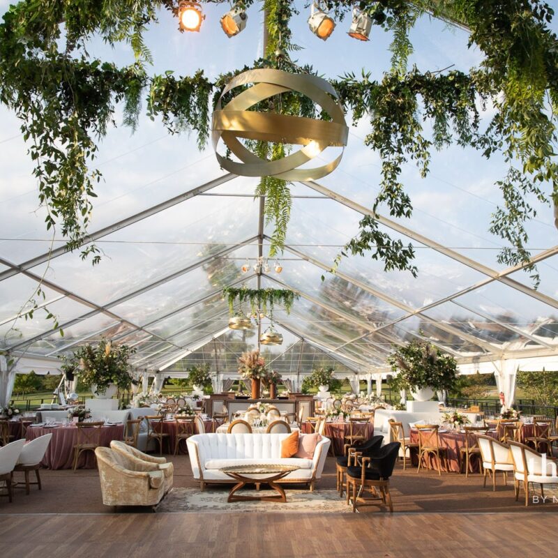 transparent event tent with golden hanging lights and greenery