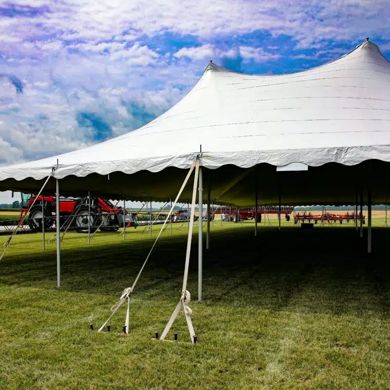 A large outdoor event tent secured with ropes and stakes, providing shade and weather protection, with agricultural equipment visible in the background.