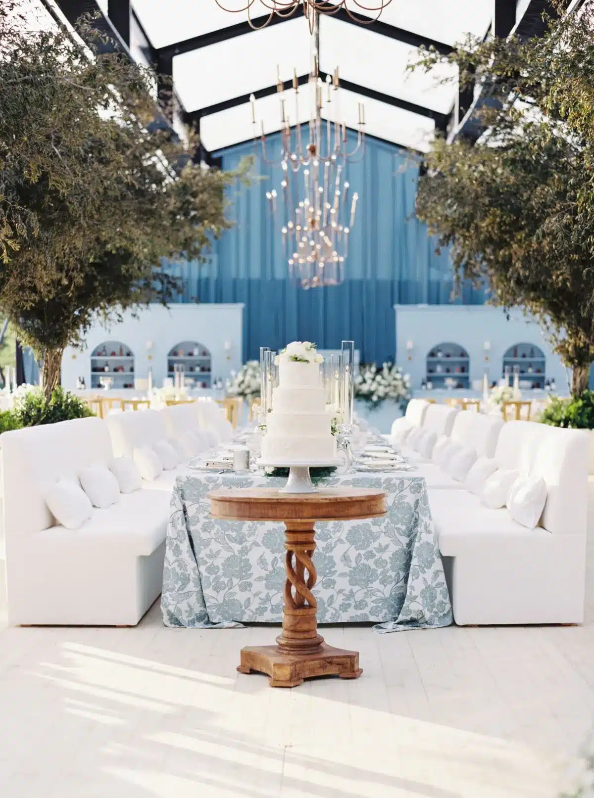 Sophisticated wedding setup inside a clear structure tent with white cushioned seating, central cake display, and blue draping for upscale event styling.
