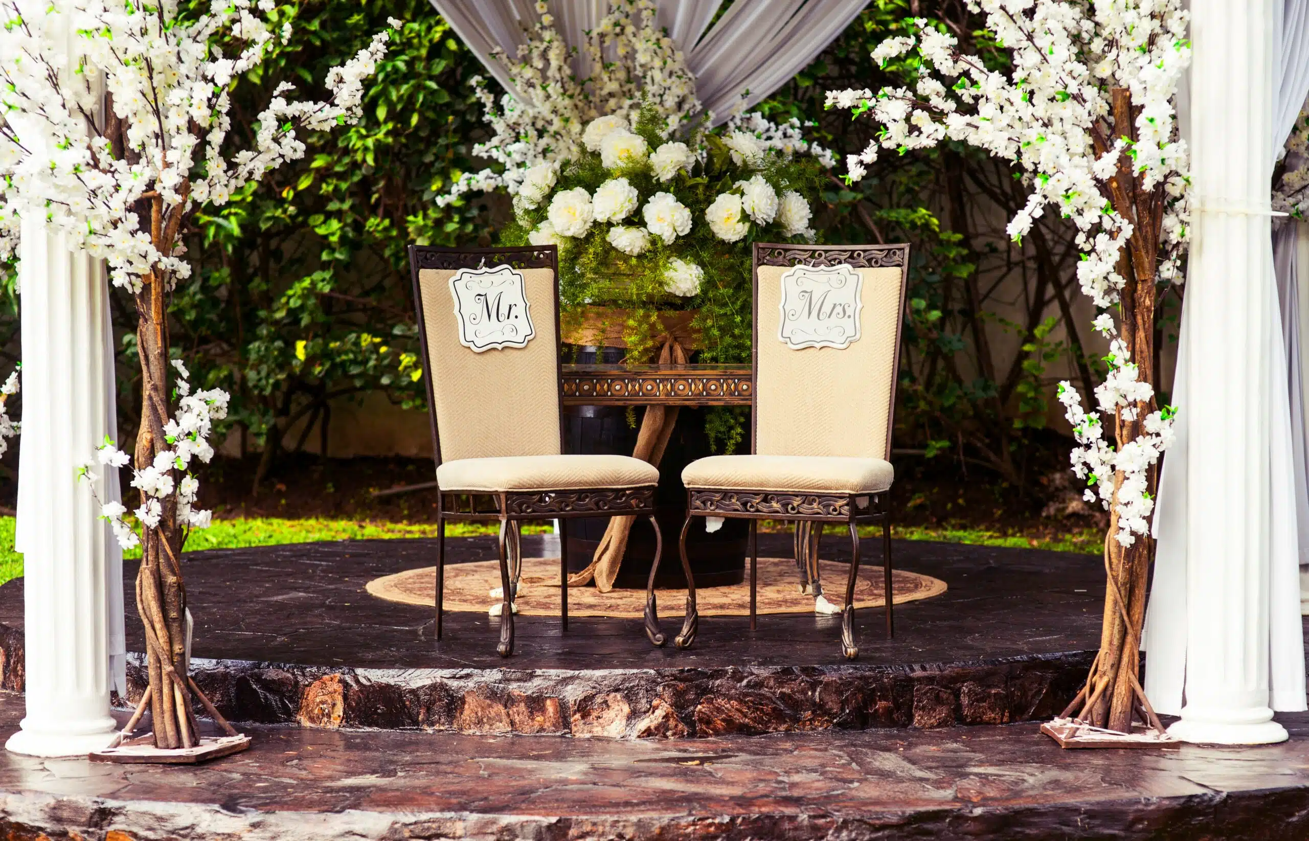 Elegant "Mr." and "Mrs." chairs sit beneath a canopy of white florals and lush greenery, blending seamlessly with the natural garden backdrop for a harmonious color palette.