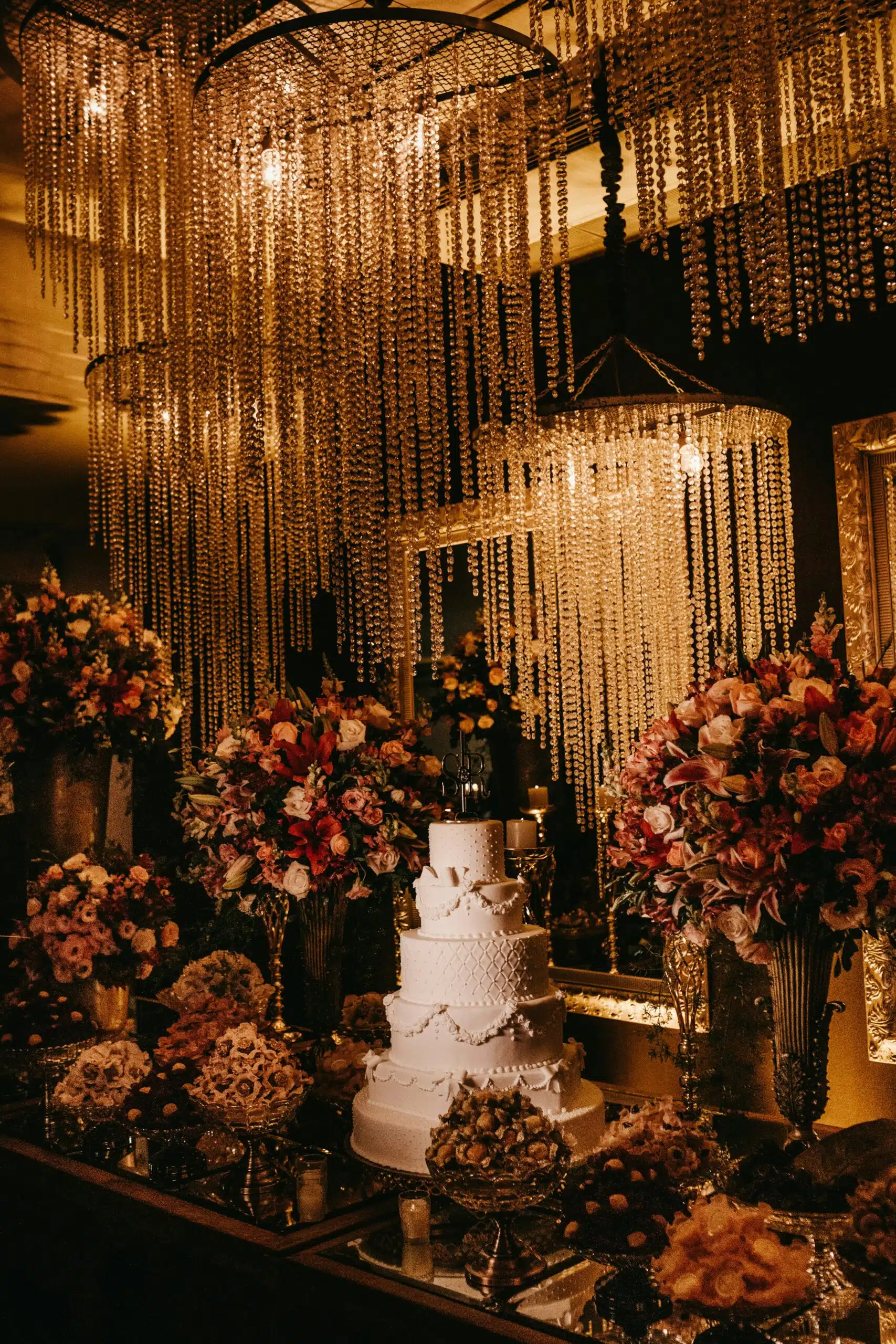 A glamorous wedding tent setup featuring cascading crystal chandeliers, lush floral arrangements, and a personalized tiered cake centerpiece.