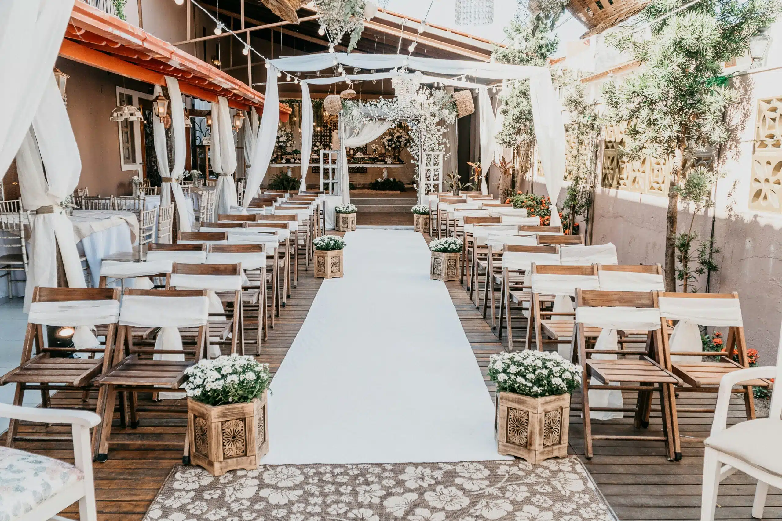 An elegantly decorated wedding aisle with wooden chairs, sheer white draping, floral arrangements, and warm ambient lighting, capturing a cohesive themed celebration.