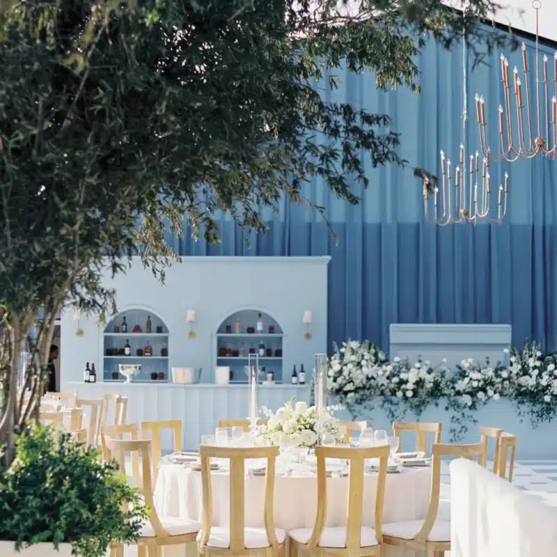 Wedding reception tables and bar area under a blue draped tent with floral decor.