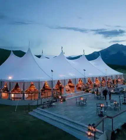 Large sailcloth tent with multiple elegant peaks and a glowing interior at dusk, demonstrating how the design’s shape and pole layout influence space planning and tent size selection.