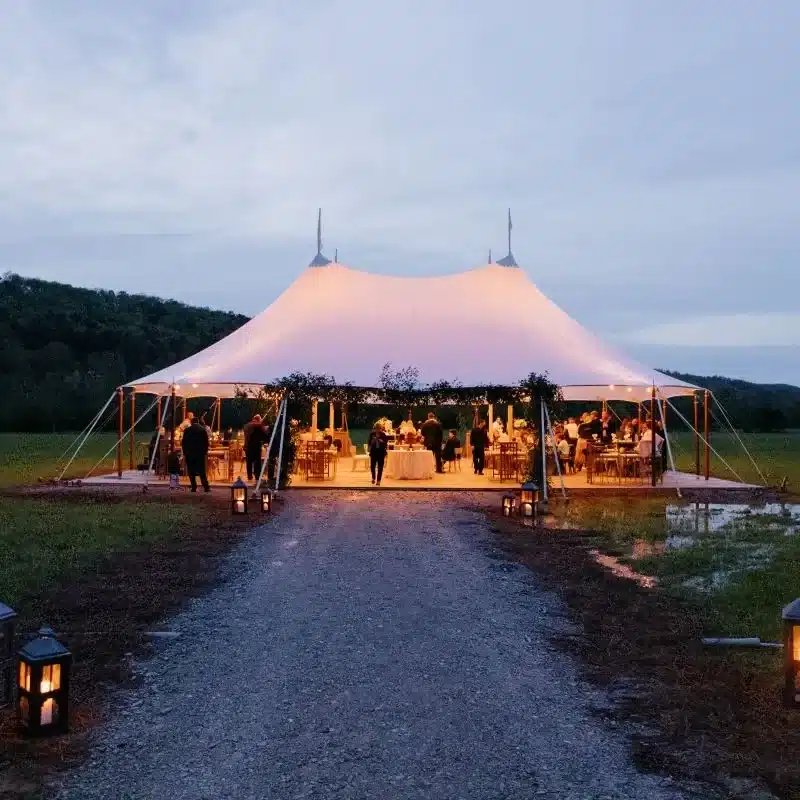 Sailcloth tent glowing warmly at dusk, set on a rural field with lanterns lining the entrance path.