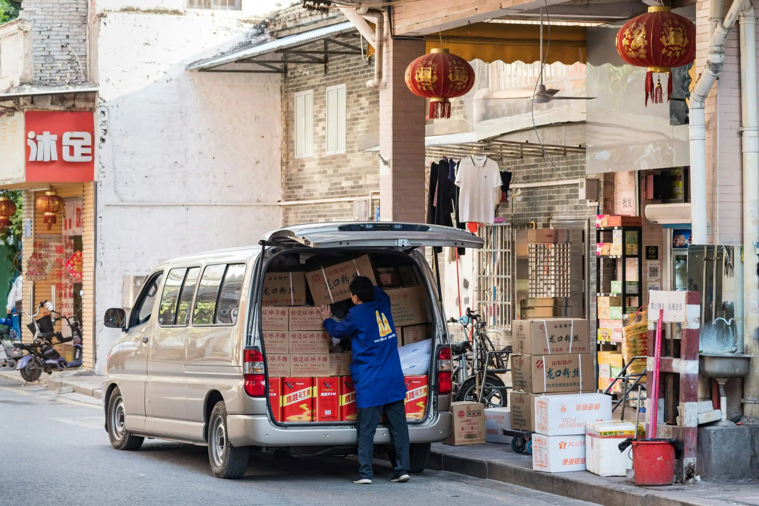 A worker unloads boxes from a packed van outside a shop, highlighting practical logistics involved in transporting and handling tent equipment for events.