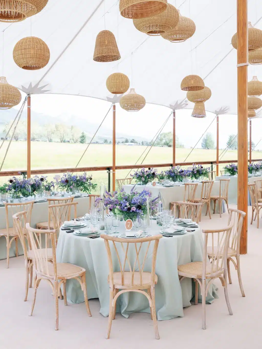 Round tables with lilac floral centerpieces under a century tent with woven pendant lighting.