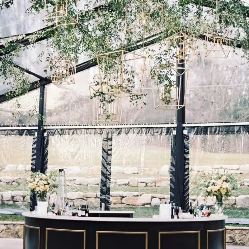 Waterproof clear-top tent featuring geometric pendant lights, greenery, and a modern black bar.