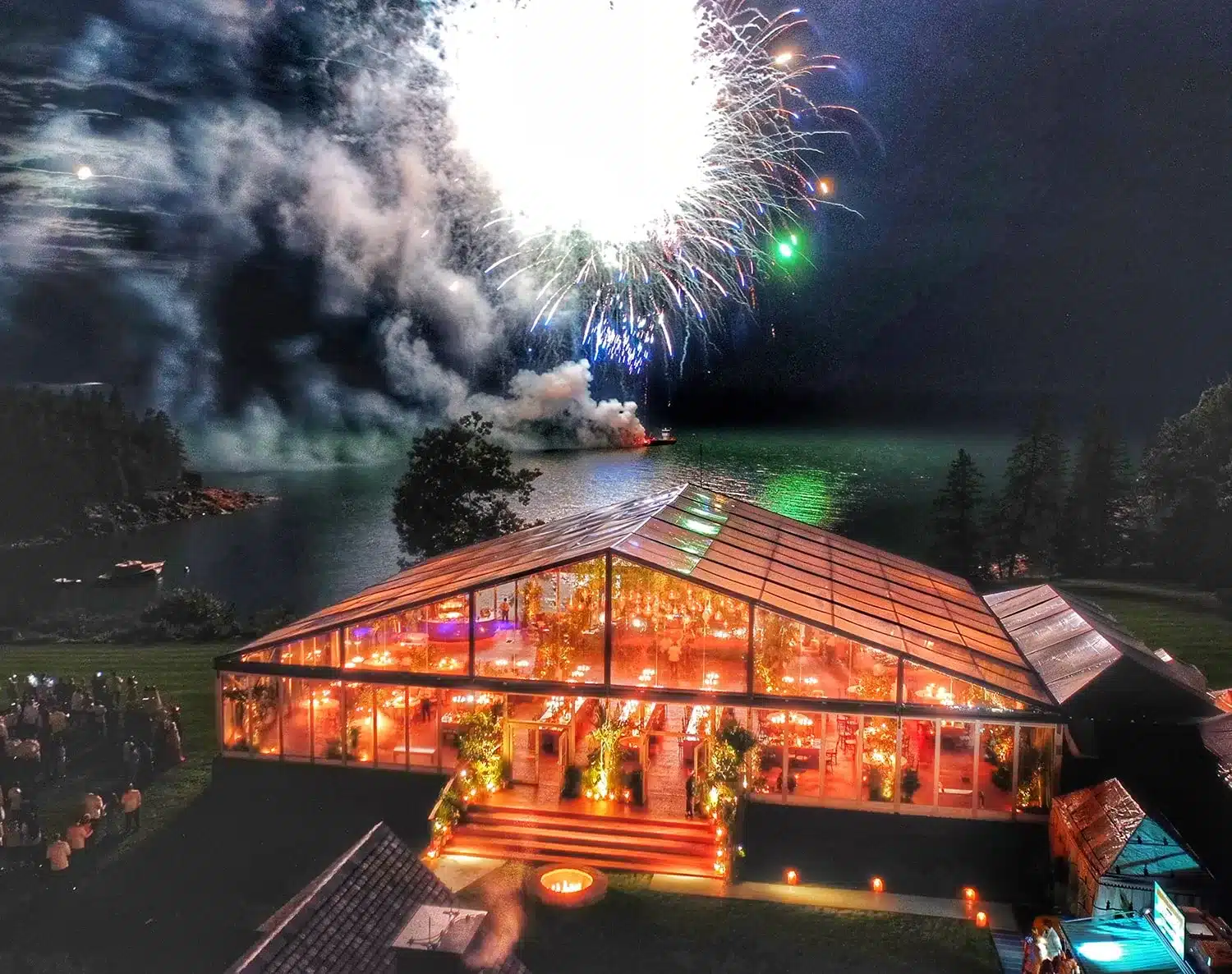 Illuminated clear event tent by a lake with fireworks in the background