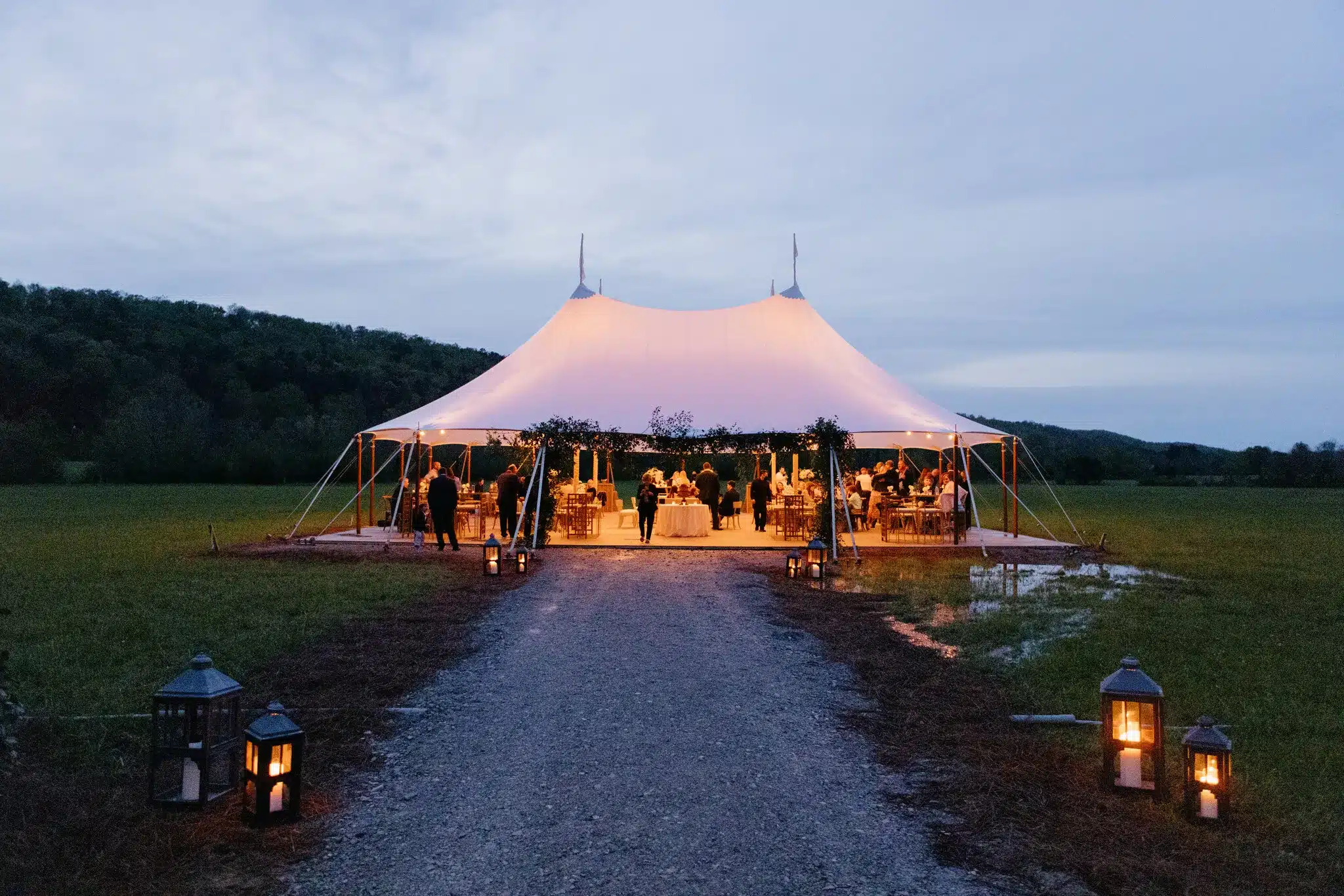 A glowing sailcloth tent set up on damp ground at dusk, emphasizing the need for elevated flooring, proper drainage, and secure anchoring to prevent weather-related damage.
