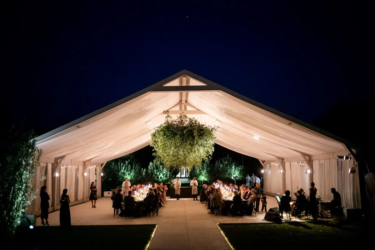 Frame-style tent glowing at night with long tables and a large greenery chandelier