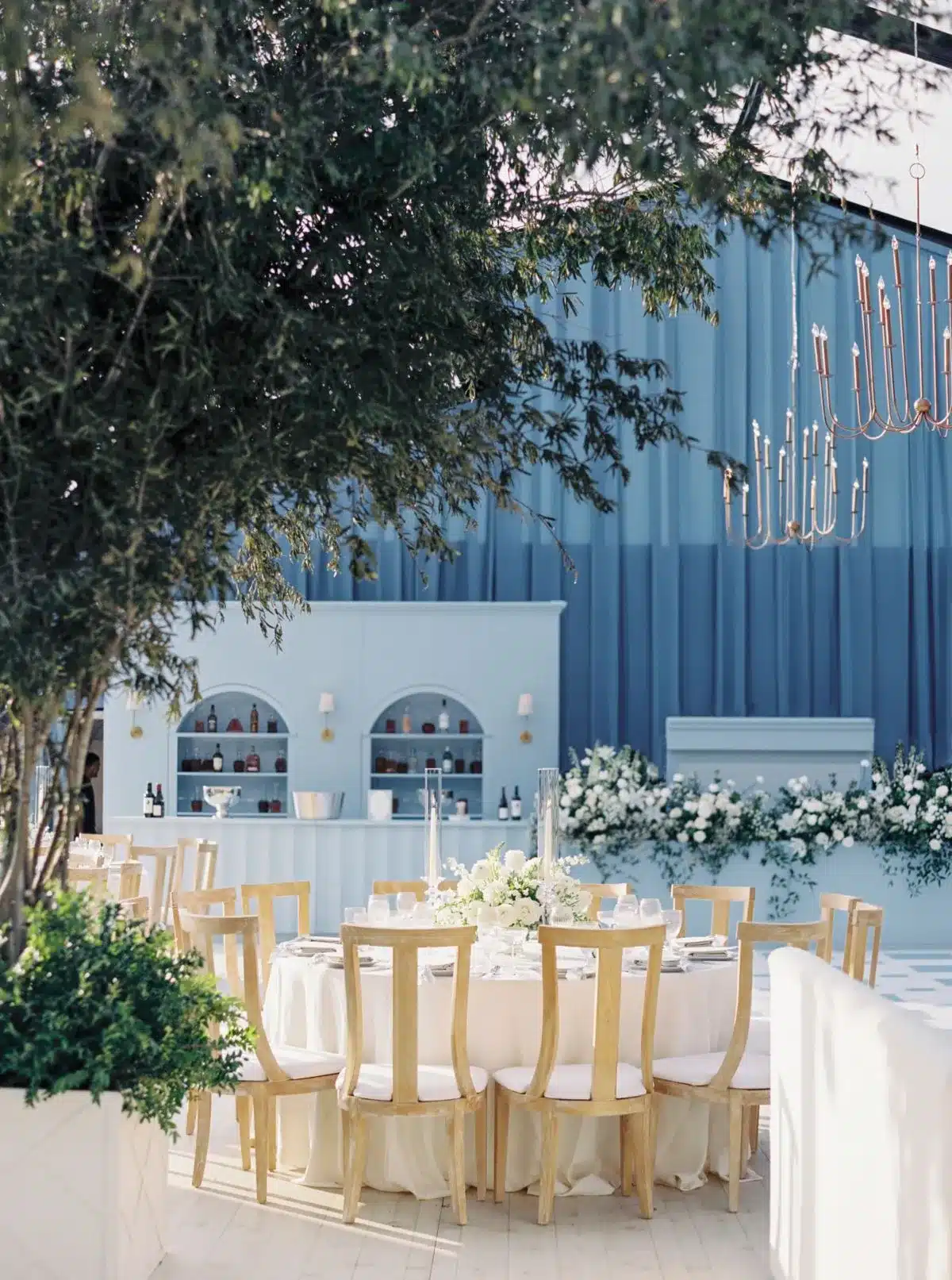 Outdoor dining setup with a white bar, round tables, and blue drapery, framed by trees and greenery.
