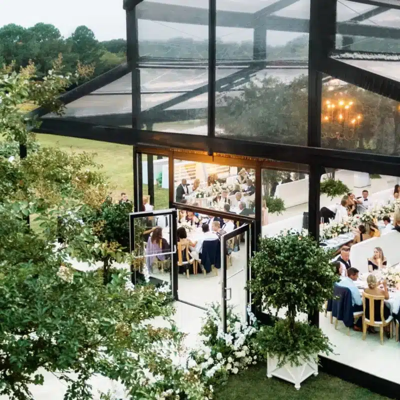 Clear tent surrounded by greenery with guests dining at long tables.