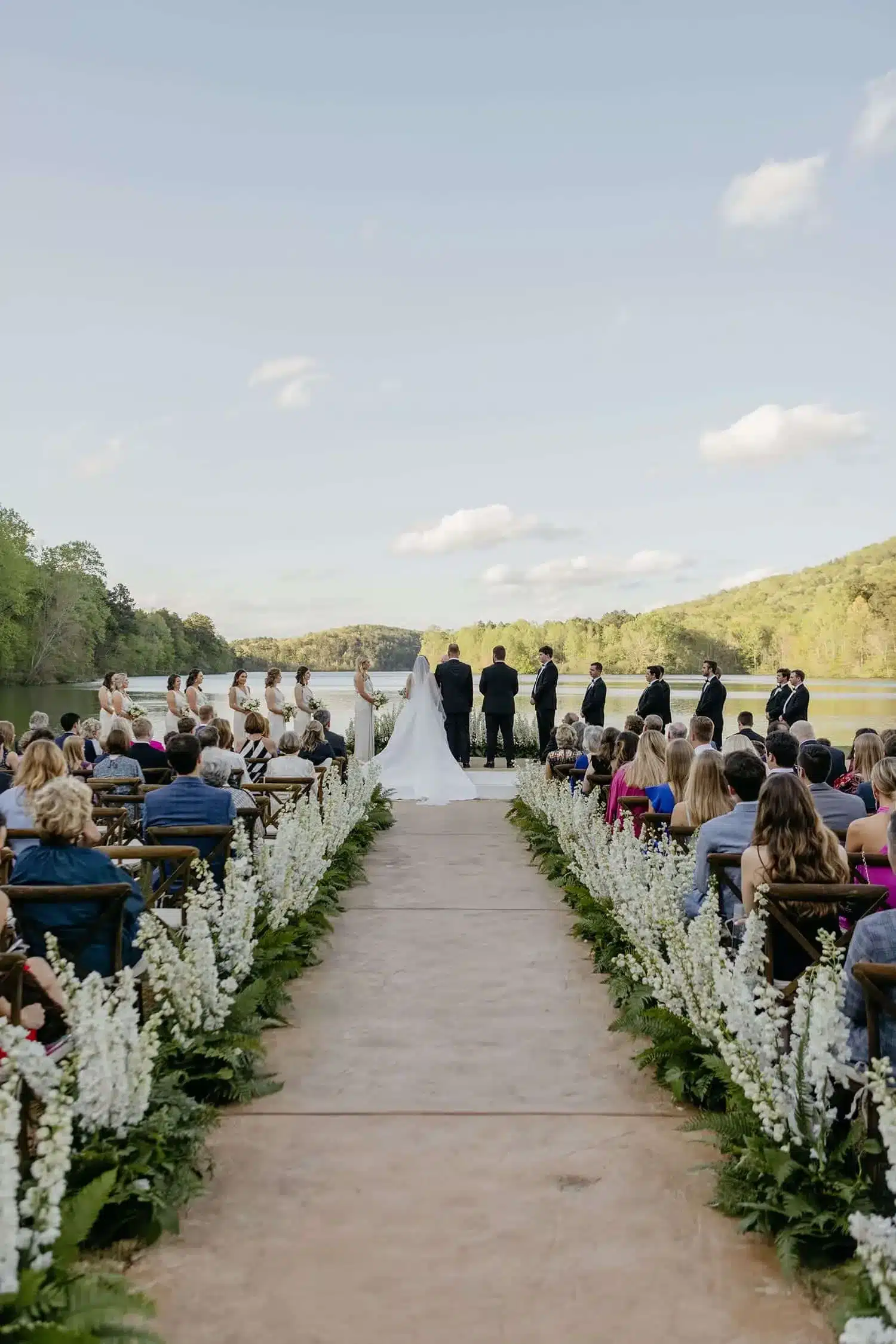 Outdoor wedding aisle lined with white floral arrangements by a lakeside.