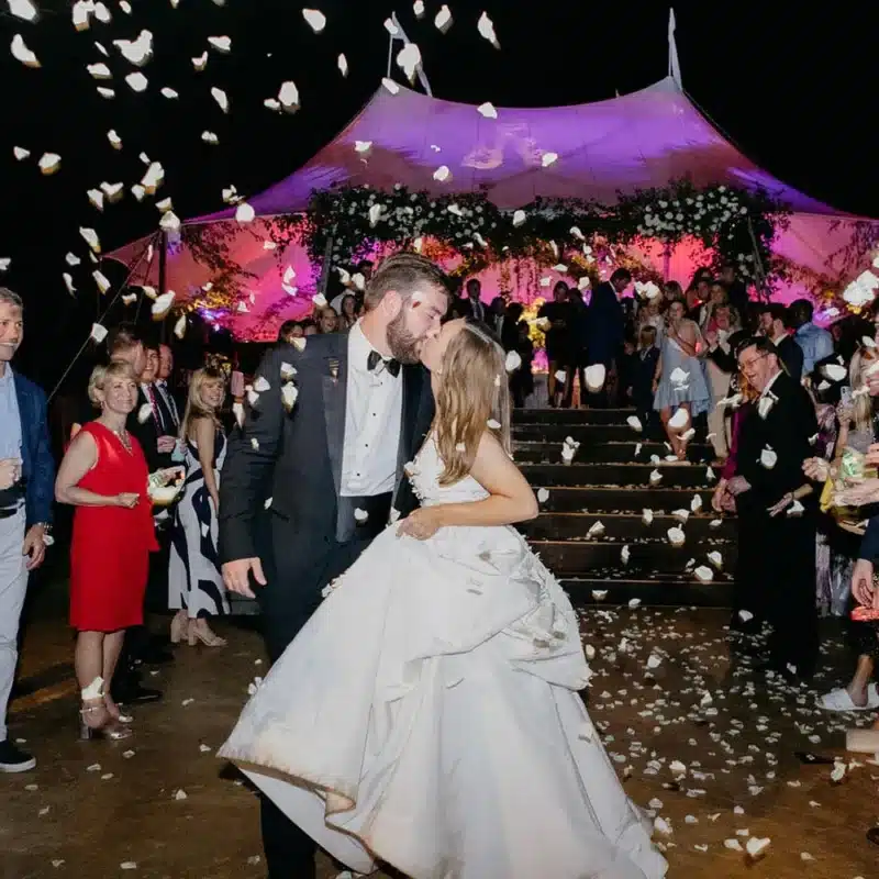 Newlywed couple kissing under a sailcloth tent with floral décor and guests celebrating.