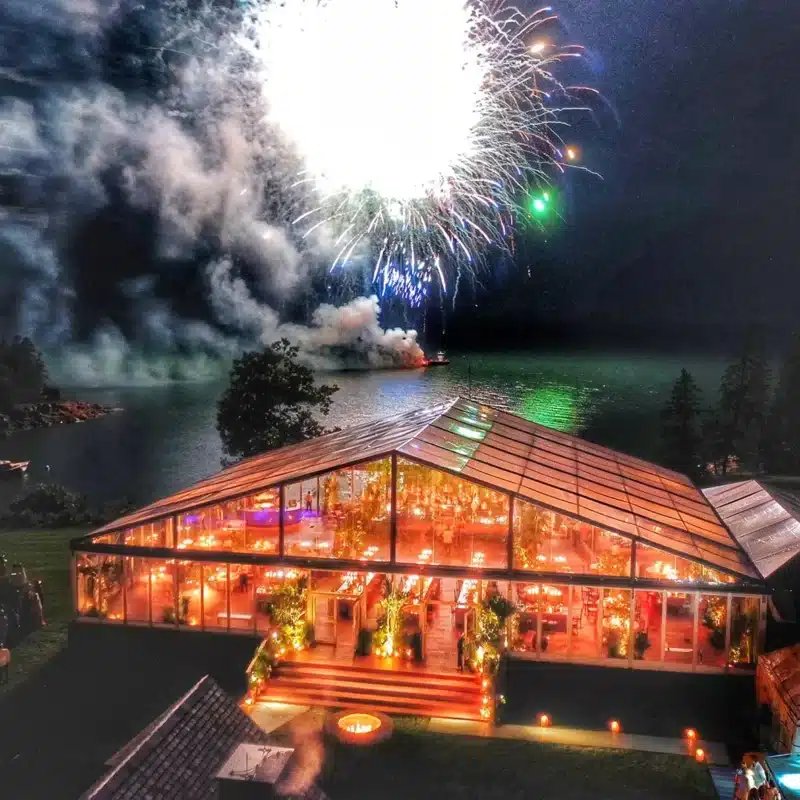 Clear-top event tent glowing at night with fireworks over a lake.