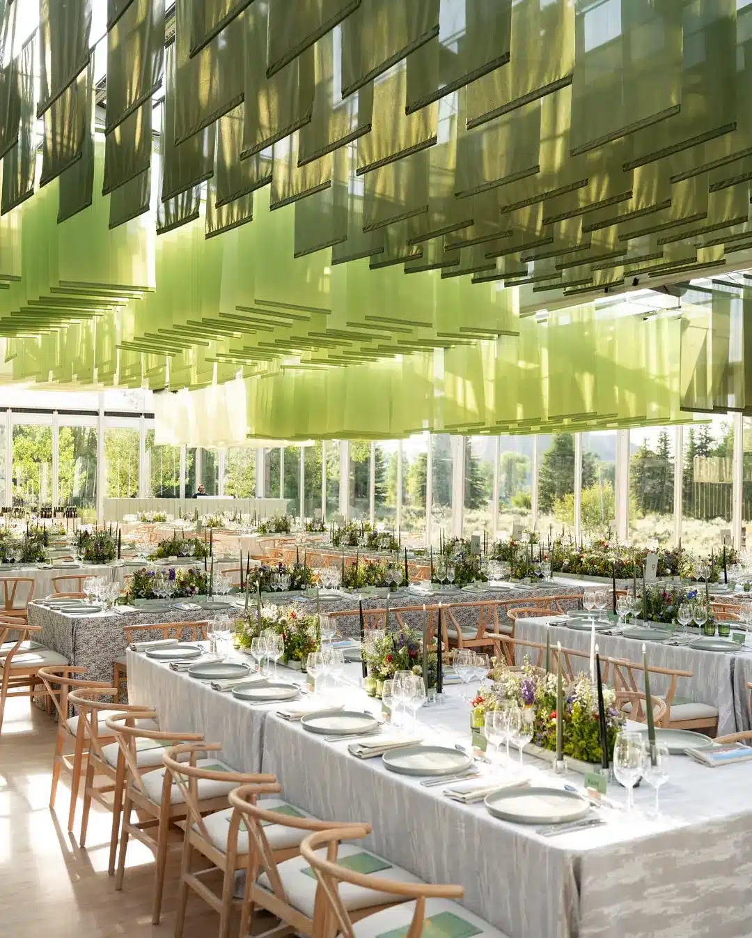 Modern glass tent with hanging green decor panels and long banquet tables set for a winter celebration