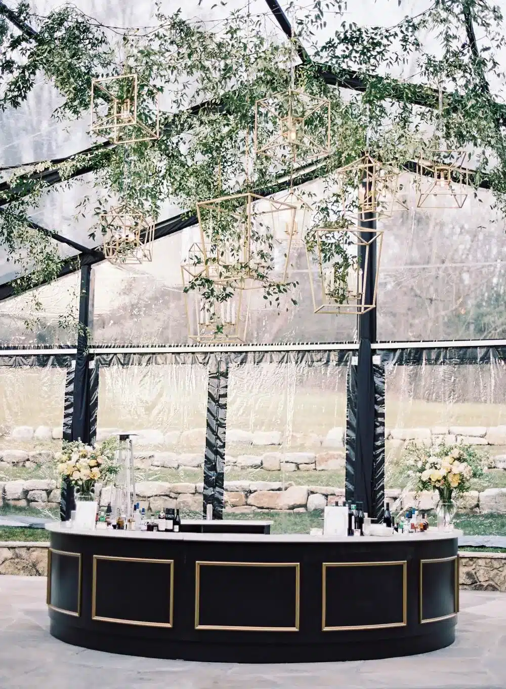 Elegant bar setup under a clear-top tent with hanging greenery and geometric gold lighting.