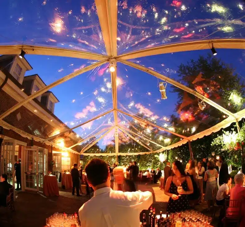 Guests mingling under a clear-top tent with reflections of fireworks