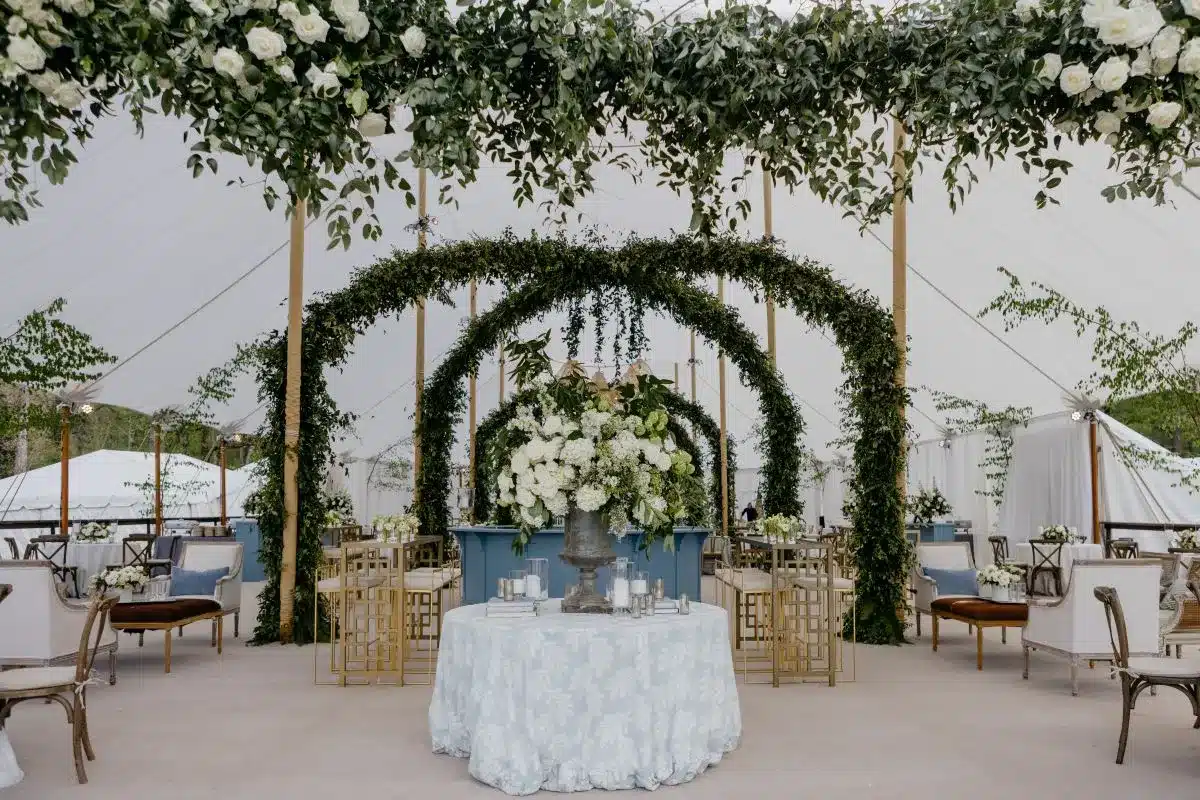 Tent lounge with pale blue linens, greenery-wrapped columns, and oversized white floral centerpiece.