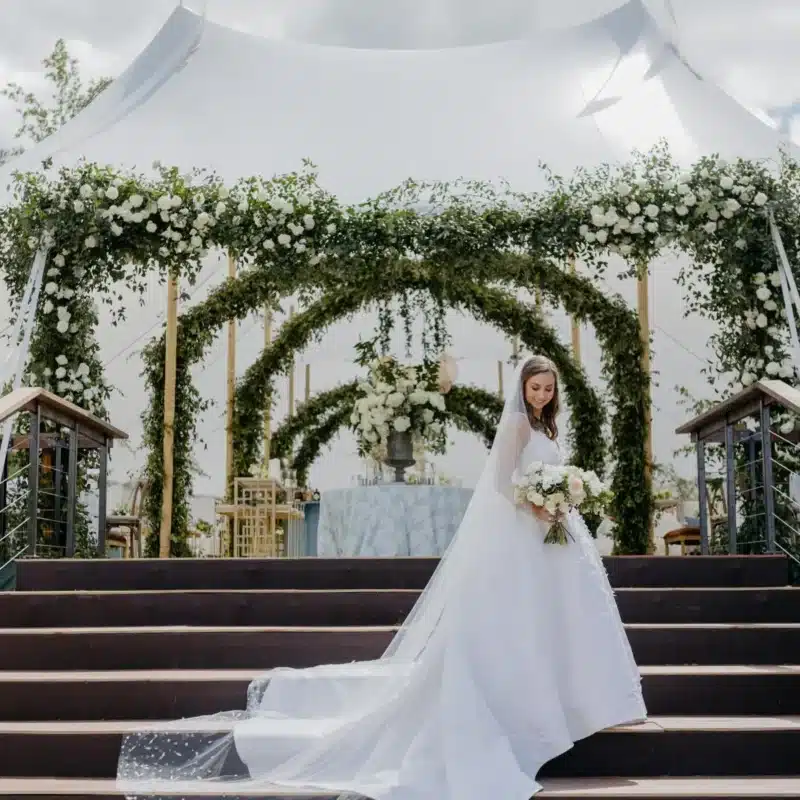 Bride in elegant white gown with veil, descending steps adorned with greenery and flowers, under a luxurious wedding tent, showcasing outdoor event design.