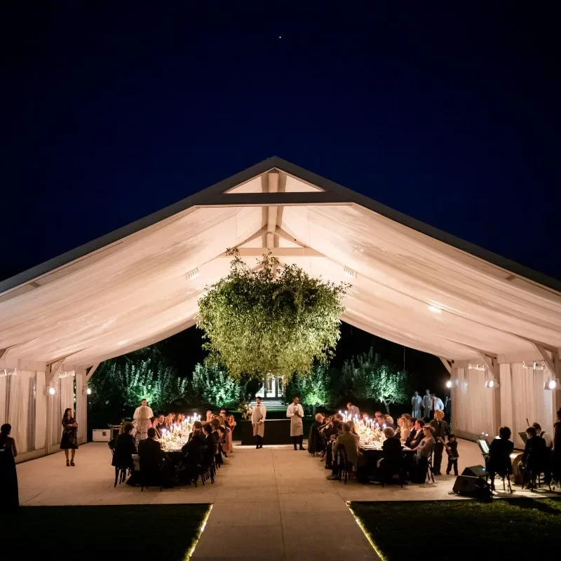 Elegant nighttime dinner party under a draped white backyard tent with long tables, warm lighting, and greenery.