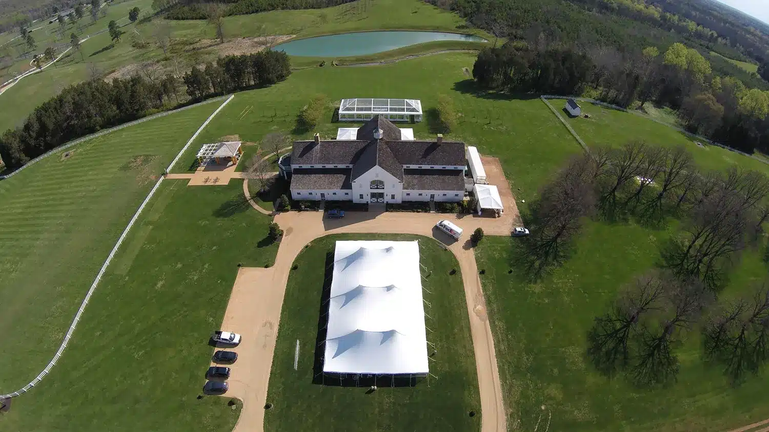 Aerial view of a spacious outdoor event venue featuring a large white tent, well-maintained green lawns, and a picturesque building, illustrating effective rain contingency planning for events by Skyline Tent Company.