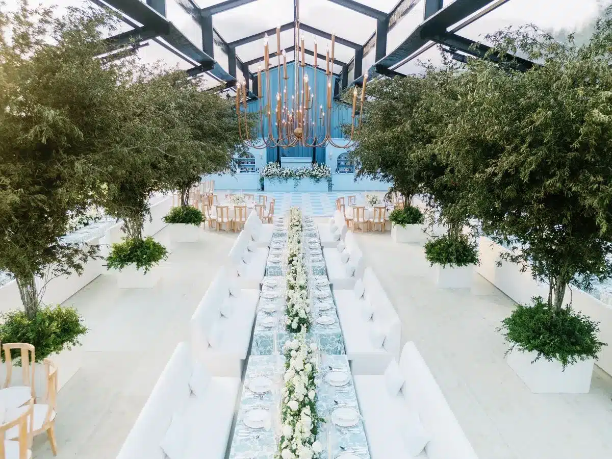 Spacious wedding tent interior with long banquet table, greenery-lined aisle, and chandeliers
