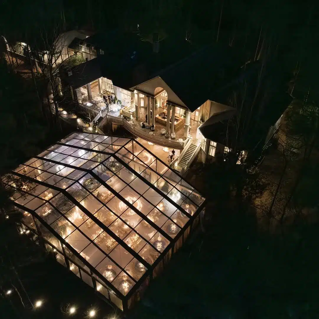 Aerial view of a clear-top wedding tent glowing with chandeliers at night