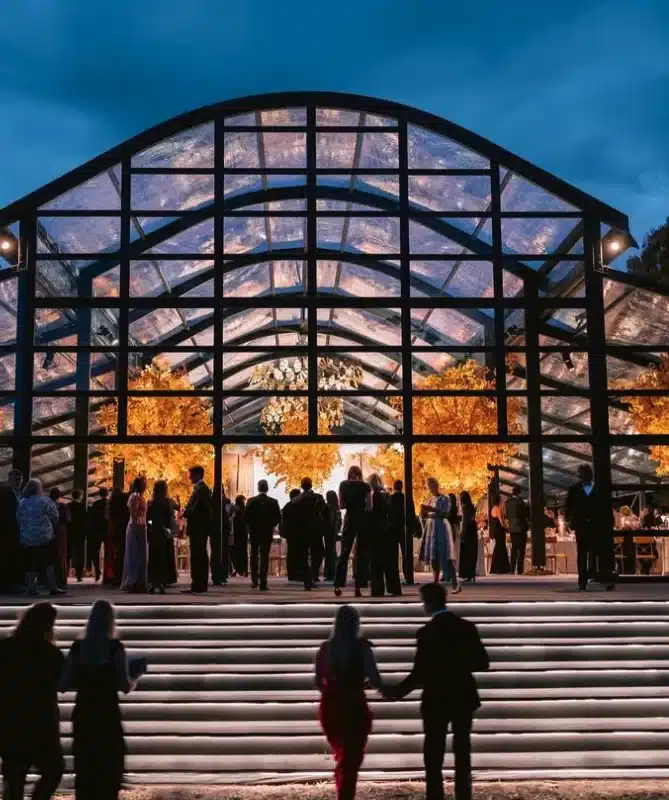 Elegant event venue with glass structure, guests mingling, vibrant autumn foliage, and warm lighting, ideal for weddings and corporate gatherings.