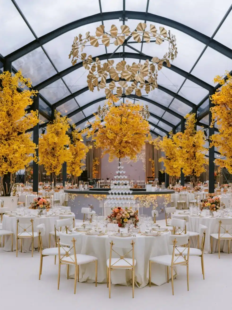 Clear-span tent with dramatic gold chandeliers and illuminated yellow trees over an elegant reception layout.