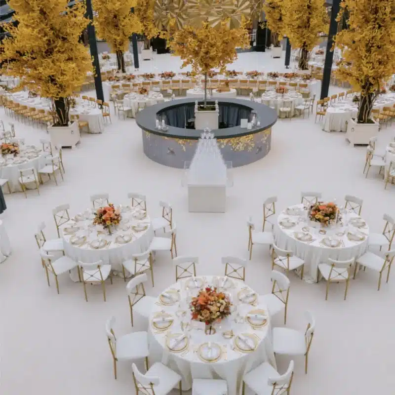 Elegant indoor event space with round tables adorned with floral centerpieces, gold accents, and white tablecloths, surrounded by vibrant yellow trees and a central bar area, ideal for luxury tent rentals and outdoor celebrations.