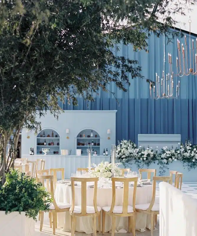 ### Key Elements

#### Main Subjects
- Round dining table with elegant setting
- Bar area in the background

#### Notable Features
- Light blue backdrop and decor
- Floral arrangements on the table and around the bar
- Wooden chairs surrounding the table
- Chandelier hanging above

#### Objects
- White tablecloth and centerpieces
- Glassware and cutlery on the table
- Decorative elements on the bar

#### Text or Symbols
- No visible text or symbols

### Overall Mood
The scene conveys a serene and sophisticated atmosphere, ideal for an elegant gathering or celebration. The combination of blue tones, floral accents, and careful arrangement suggests a tasteful, inviting space.