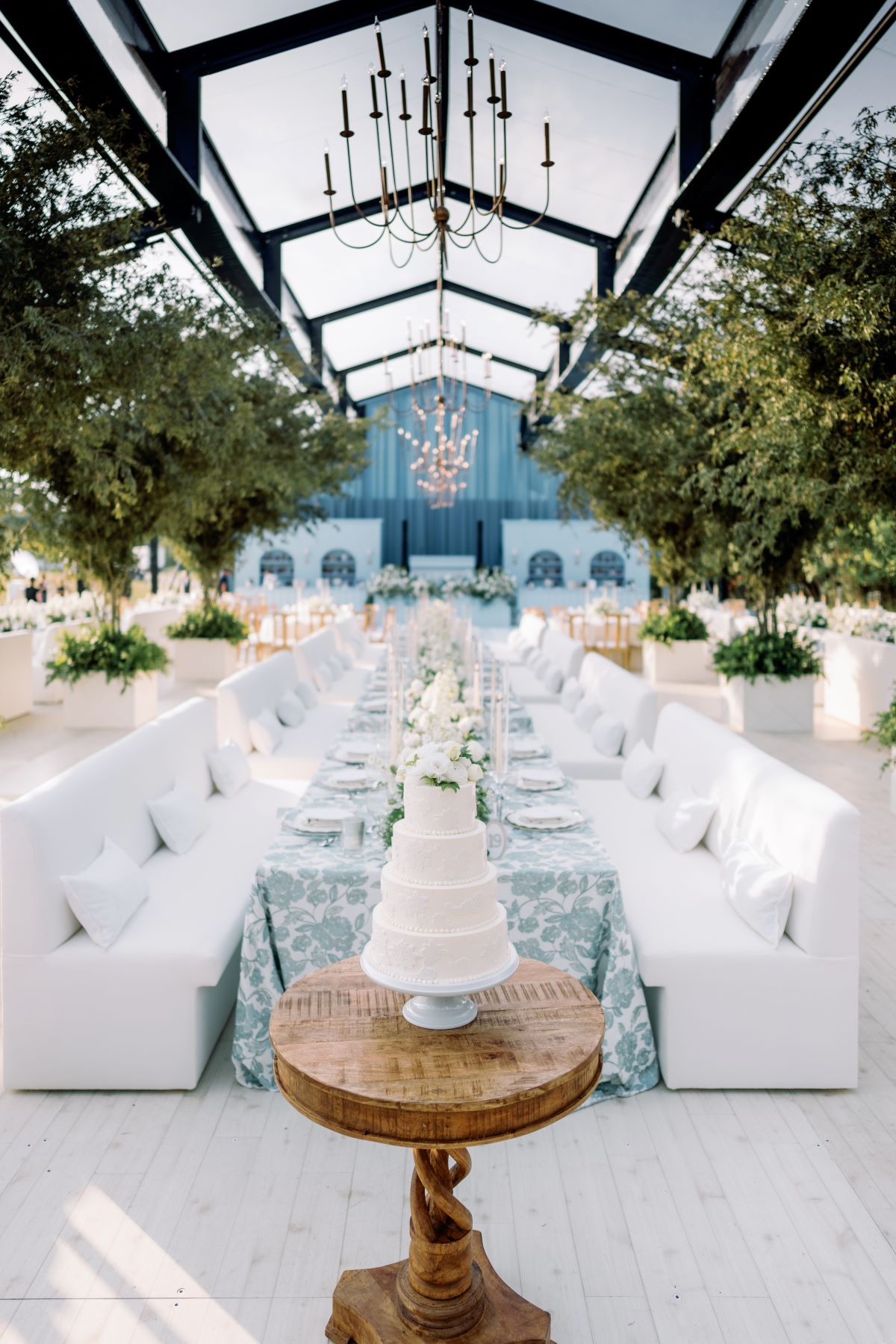 Long banquet table with white seating lined under a tent, framed by greenery and chandeliers.