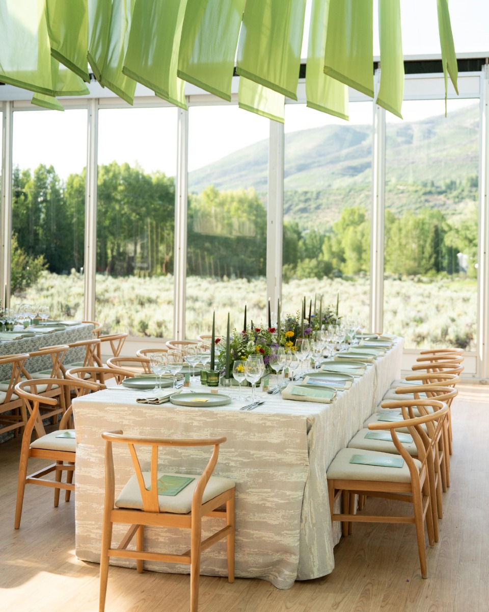 Sunlit dining table inside a glass tent with soft green drapery and scenic outdoor views.