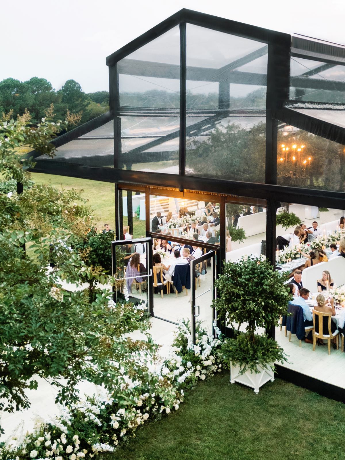 Elevated view into a glass-walled tent with guests dining under chandeliers.