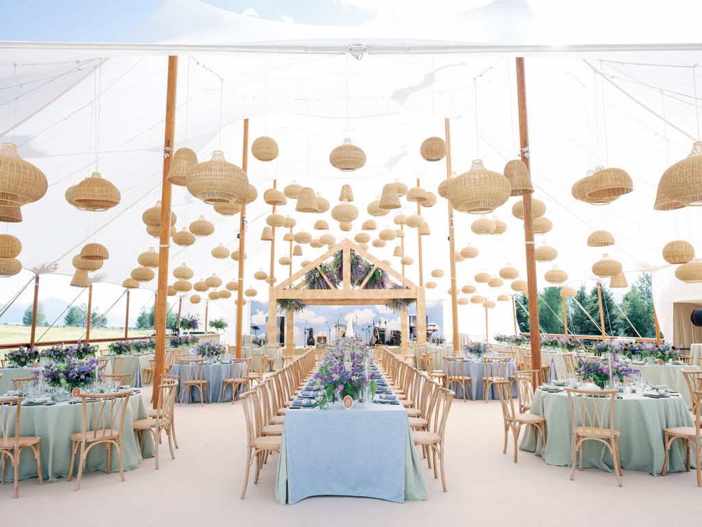 Mixed seating layout with long and round tables under a sailcloth tent filled with hanging basket lights.